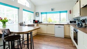 Una cocina moderna con suelo de madera, armarios blancos, zona de comedor con mesa y sillas, y ventanas amplias con persianas azules.
