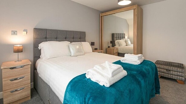 Quarto de hotel moderno com uma cama grande, uma mesa de cabeceira com abajur, um criado-mudo de madeira, um espelho e um cobertor azul-turquesa.