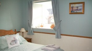 A bedroom with a bed, a window with curtains, a bedside table with a lamp, and a painting on the wall. - Swallow Cottage (Alnwick)