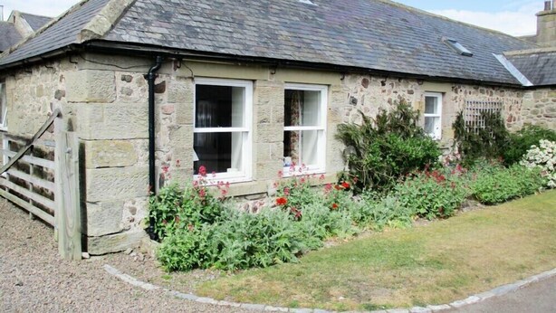 A stone cottage with a sloped roof, a chimney, and a small garden with flowers and shrubs. - Swallow Cottage (Alnwick)