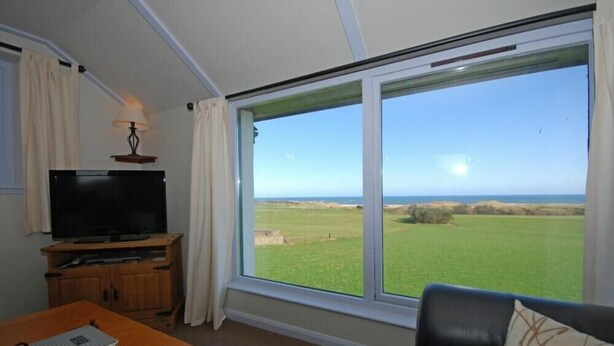 Habitación con un ventanal grande que ofrece vista a un campo verde y al mar. Hay un televisor sobre un mueble de madera y una mesa con una libreta.