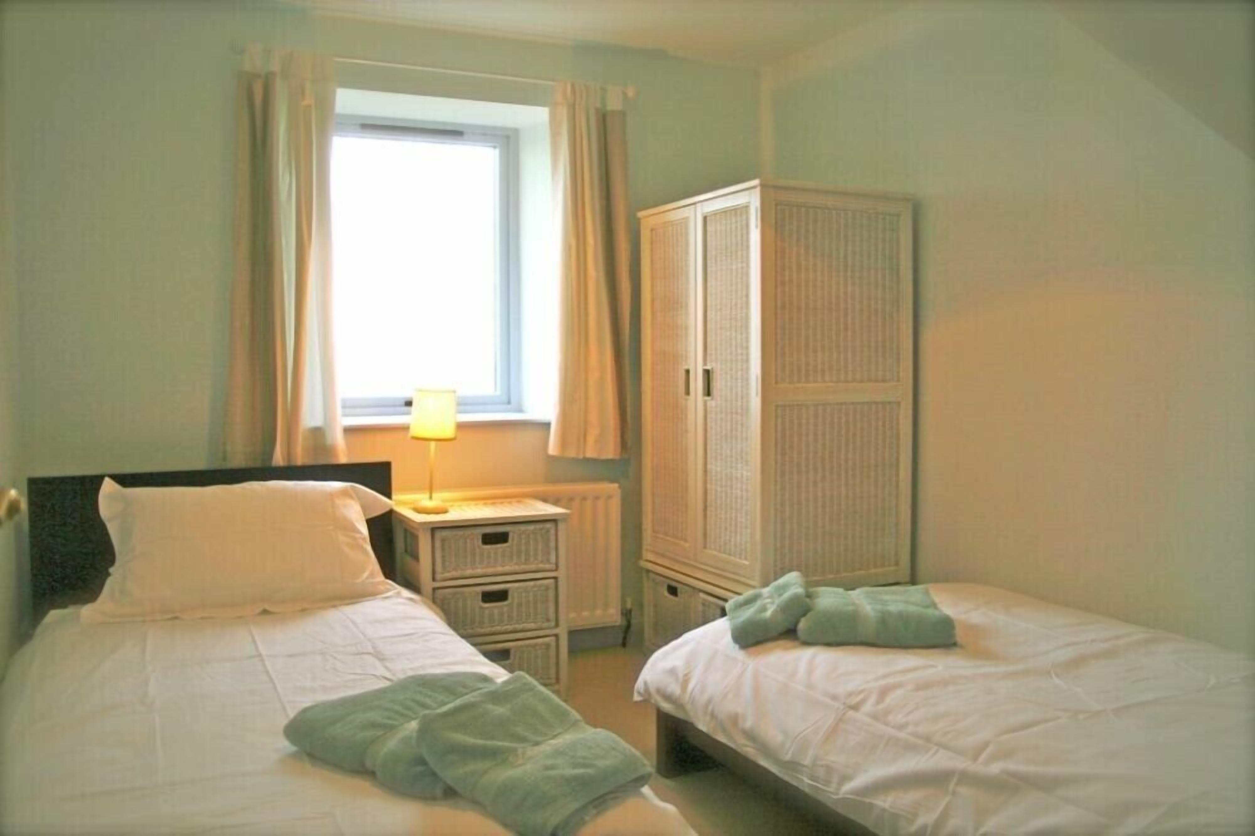 A bedroom with two beds, a window with curtains, a bedside lamp, a wardrobe, and a radiator.