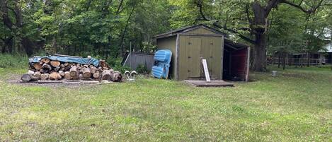 Une zone herbeuse avec une pile de bois de chauffage, un abri de jardin et une bâche bleue.