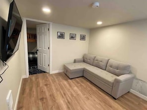 Een woonkamer met een beige hoekbank, een flatscreen televisie aan de muur en een houten vloer.