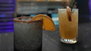 Two drinks on a bar counter with a blurred background of shelves and a person. - Aventuras Hotel (Loreto)