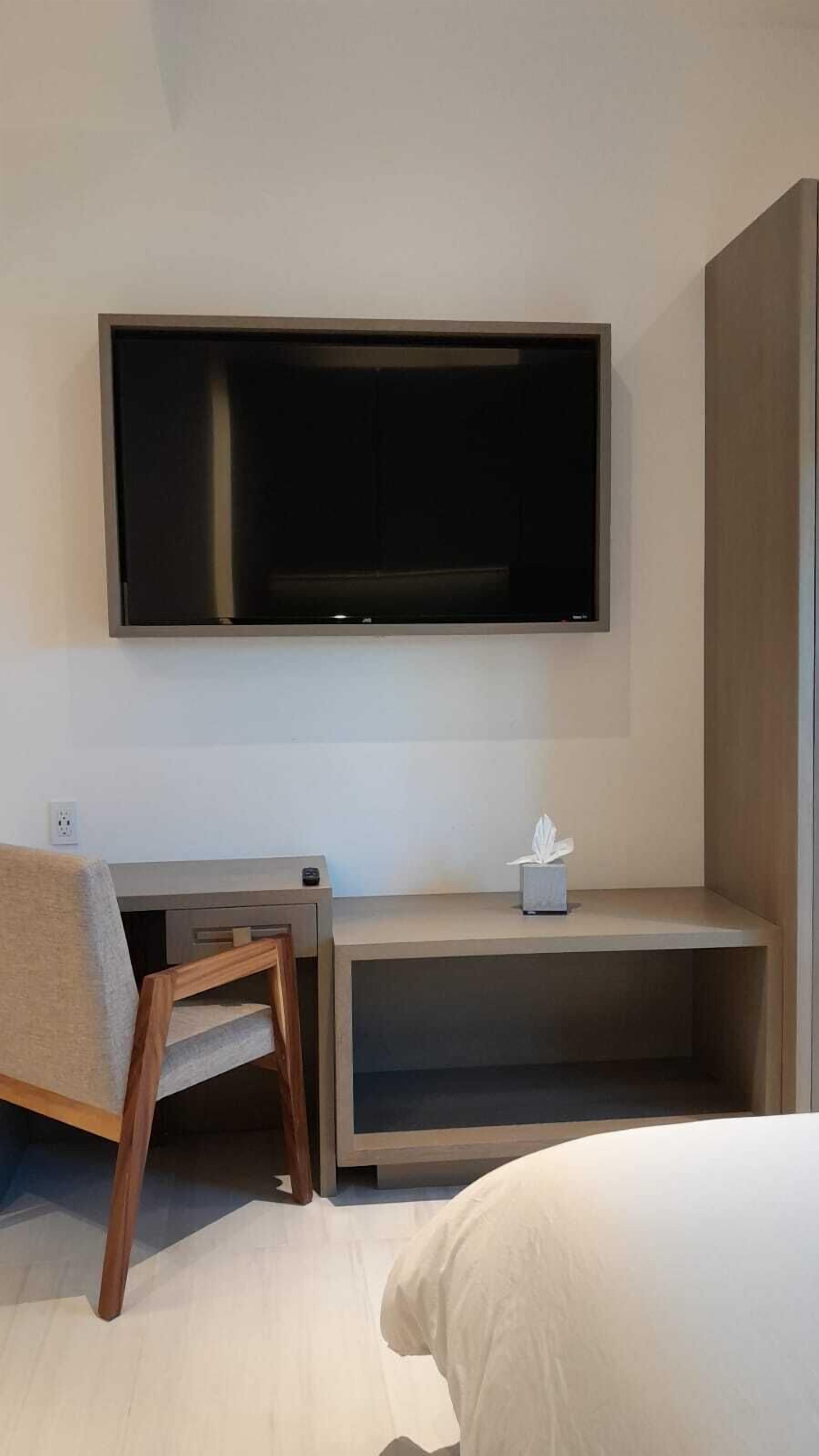 A hotel room with a bed, a desk with a chair, a flat-screen TV mounted on the wall, and a small bedside table with a tissue box.