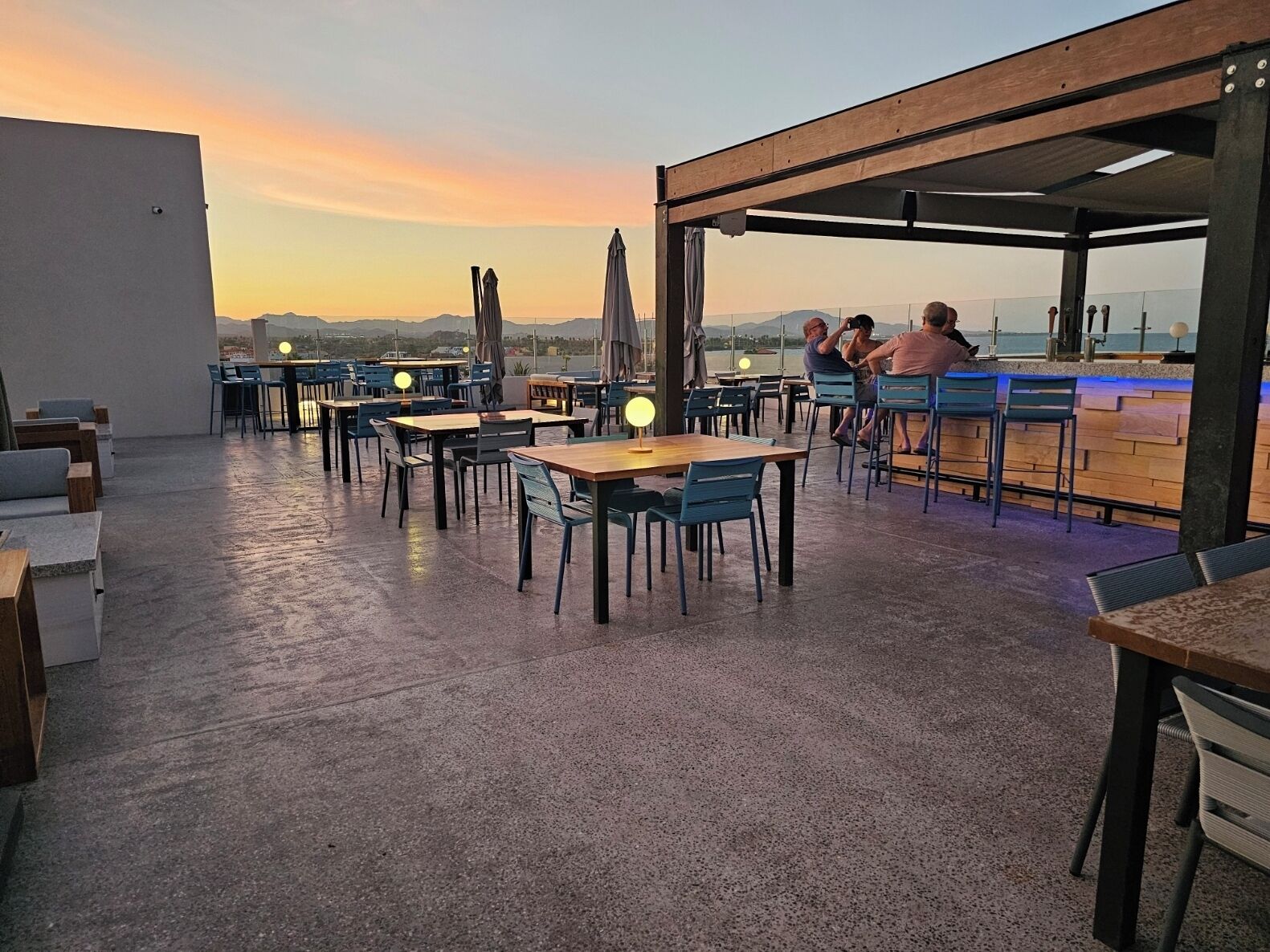 An outdoor dining area with tables and chairs, a bar, and a view of the sunset over the mountains.