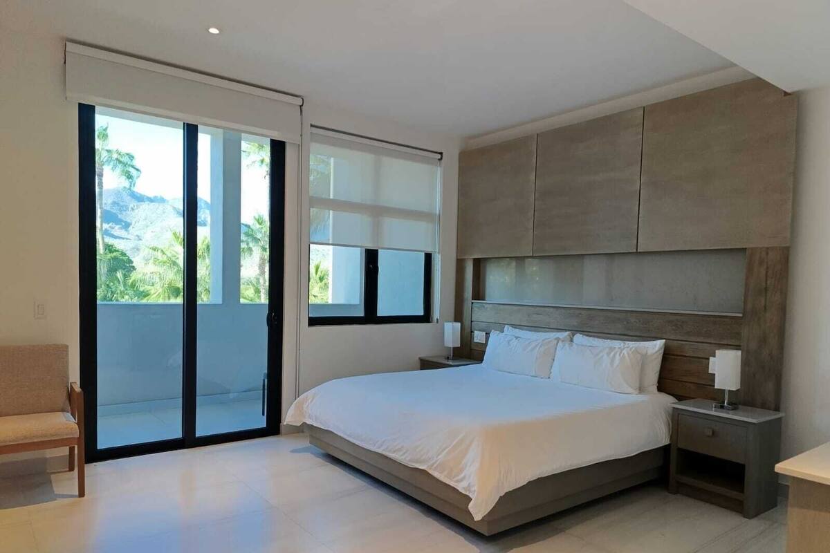 Quarto moderno com uma cama grande, mesa de cabeceira, uma cadeira e porta de vidro deslizante que dá para uma varanda.