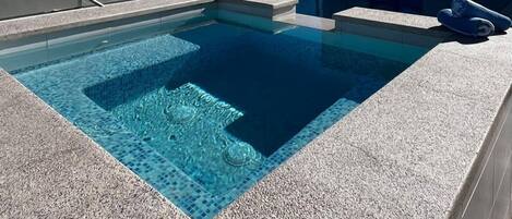 A modern pool area with clear blue water, a stone deck, and a view of the ocean and mountains in the background.