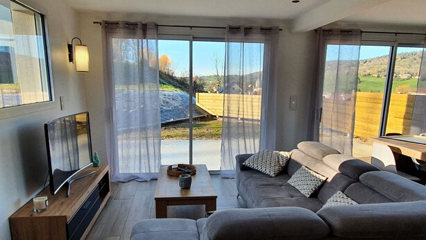 Un moderno salón con un sofá gris, una mesa de centro de madera y un televisor de pantalla plana sobre un mueble de madera. Amplias ventanas con cortinas translúcidas ofrecen vistas a un paisaje verde.
