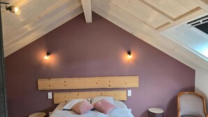 Un dormitorio con techo inclinado, cabecera de madera, cama con ropa de cama blanca, dos mesitas de noche, una silla y dos luces fijadas a la pared.