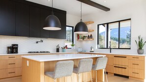 A modern kitchen with a large island, black pendant lights, wooden cabinets, and a view of the outdoors. - Mountain View Home w/ Gym, Sauna & Cold Plunge (Squamish)