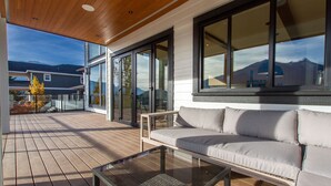 A modern outdoor deck with a glass door leading to a balcony, a sofa, and a glass coffee table. - Mountain View Home w/ Gym, Sauna & Cold Plunge (Squamish)