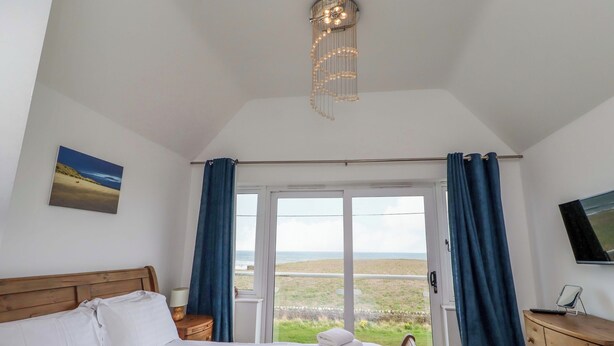 A bedroom with a bed, a bedside table, a lamp, a TV on the wall, and a view of the outdoors. - Farne Cottage (Chathill)
