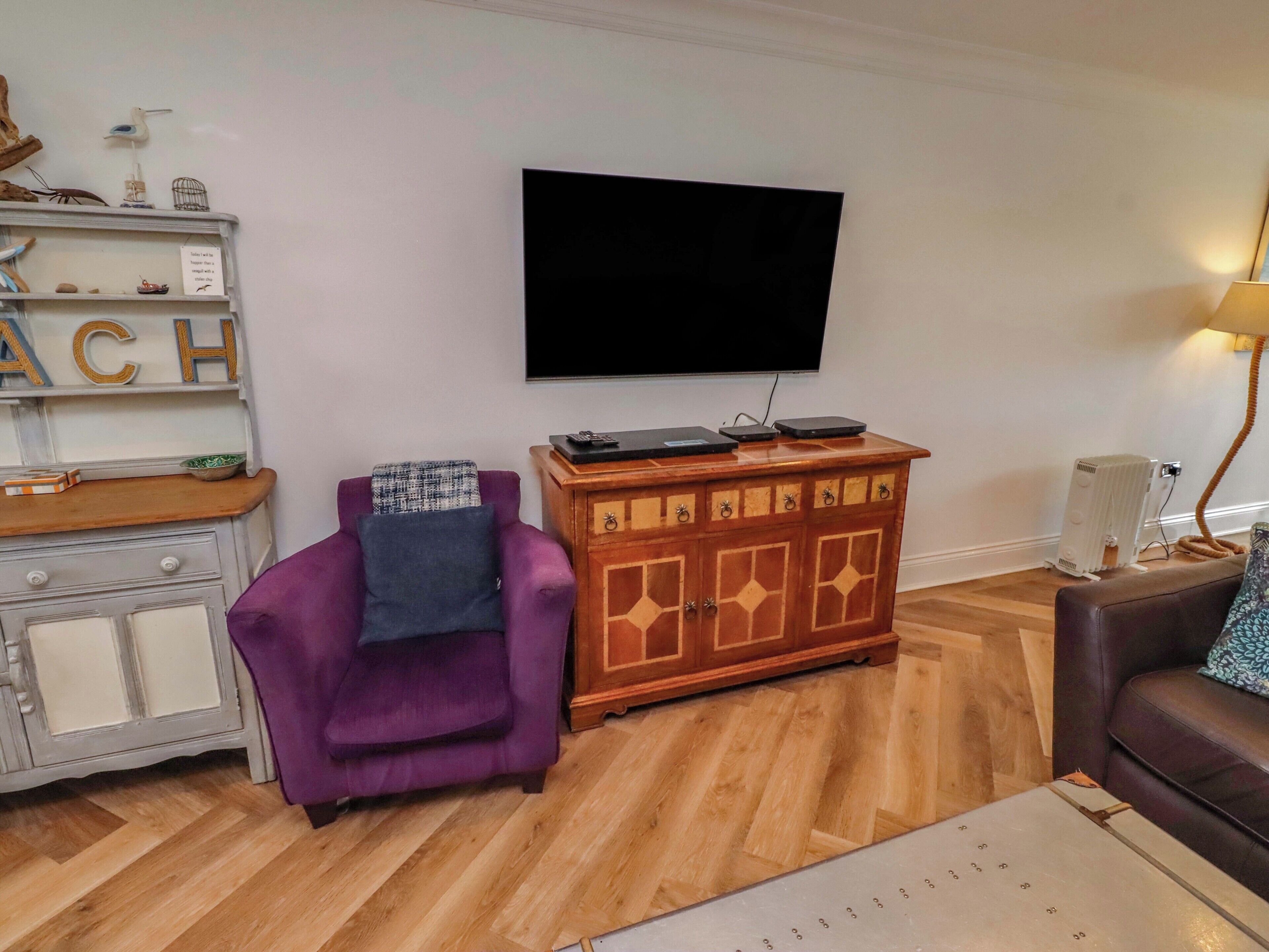 A living room with a flat-screen TV mounted on the wall, a purple armchair, a wooden cabinet, and a brown leather sofa.