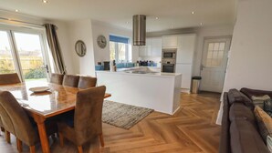 A modern kitchen with a central island, dining area with chairs, and a living room with a sofa. - Farne Cottage (Chathill)