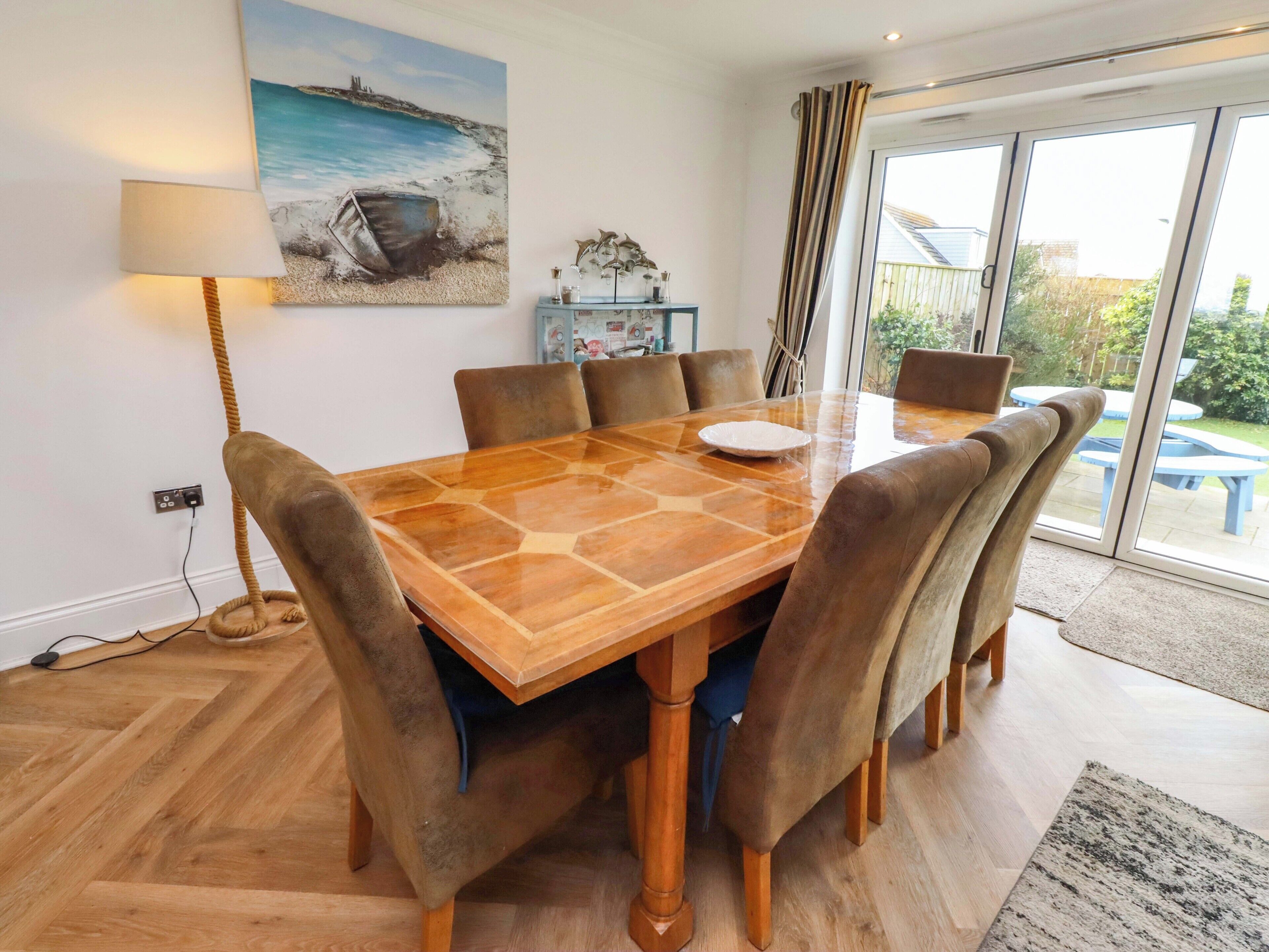 A dining area with a wooden table, six chairs, a painting of a beach scene, a lamp, and a view through a glass door.