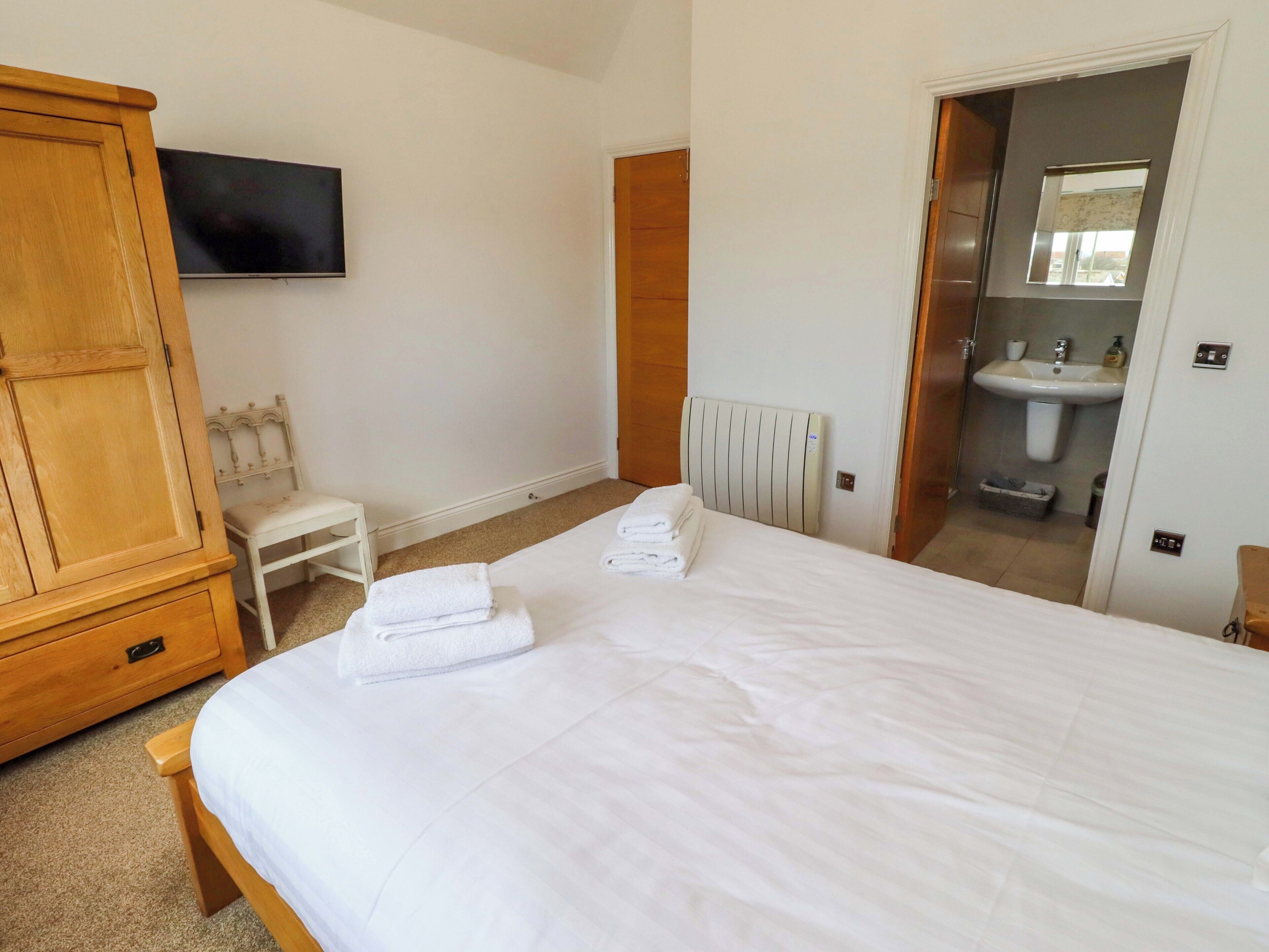 A bedroom with a bed, a wooden wardrobe, a white chair, a television, and a bathroom with a sink and mirror.