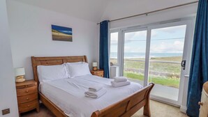 A bedroom with a wooden bed, a nightstand with a lamp, a view of the sea through a large window, and a painting on the wall. - Farne Cottage (Chathill)