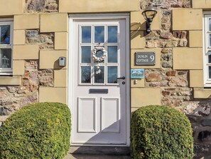 A white door with a glass panel, a stone wall, a sign reading 'DUNLIN COTTAGE 9', and two trimmed bushes.