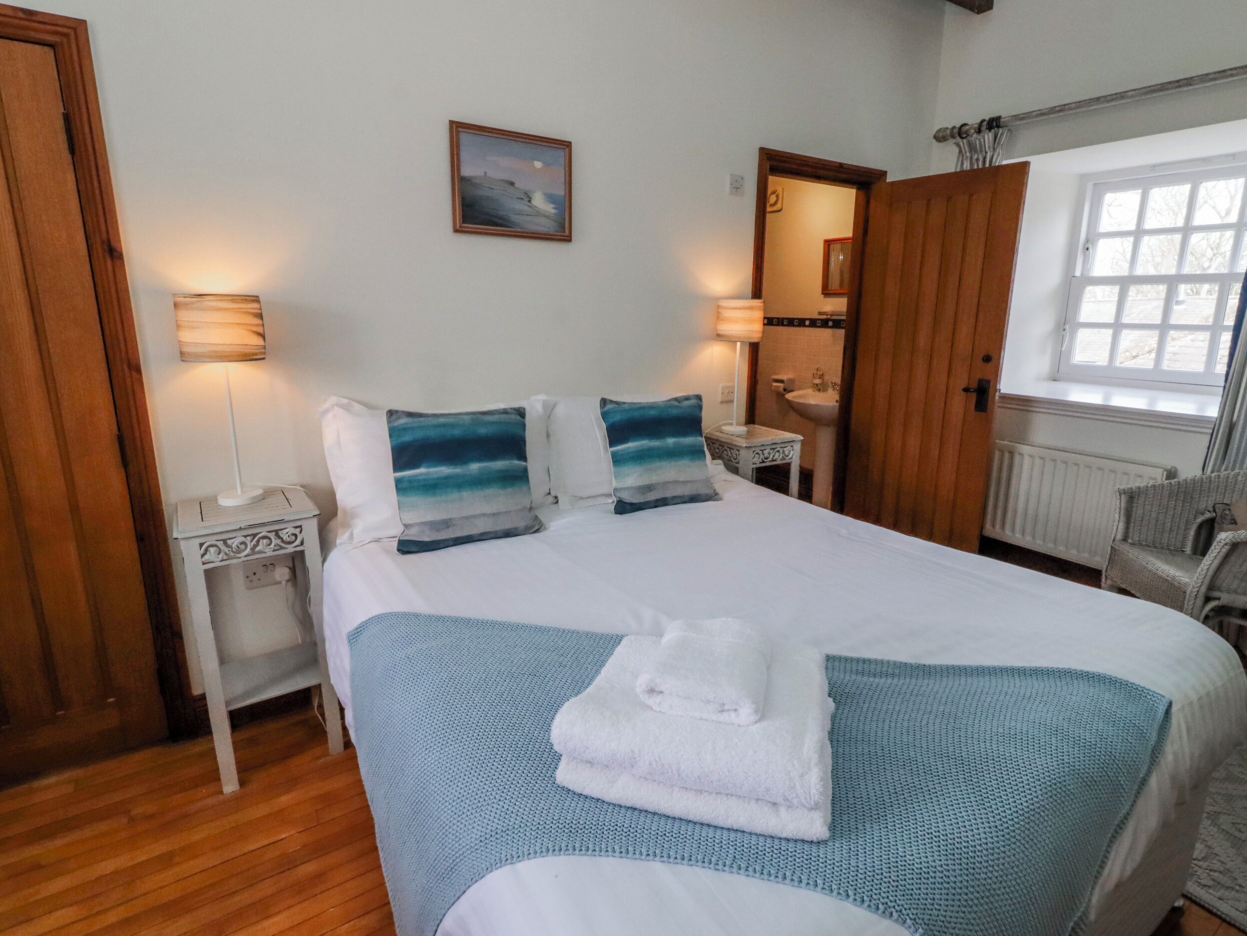 A neatly made bed with pillows, a bedside table with a lamp, a window with curtains, and a mirror.