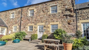 A stone building with multiple windows, a door, and a small outdoor seating area with a bench and a table. - No9 Budle Sands (Bamburgh)