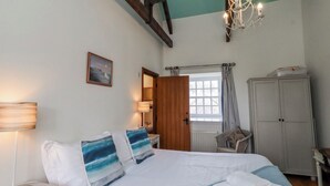 A bedroom with a bed, a nightstand, a dresser, a chair, a window with curtains, a chandelier, and a painting on the wall. - No9 Budle Sands (Bamburgh)