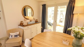 Uma sala de jantar com uma mesa redonda de madeira, cadeiras brancas, uma poltrona cor creme, um espelho, um letreiro com a palavra 'Bamburgh' e vista através de uma porta de vidro.
