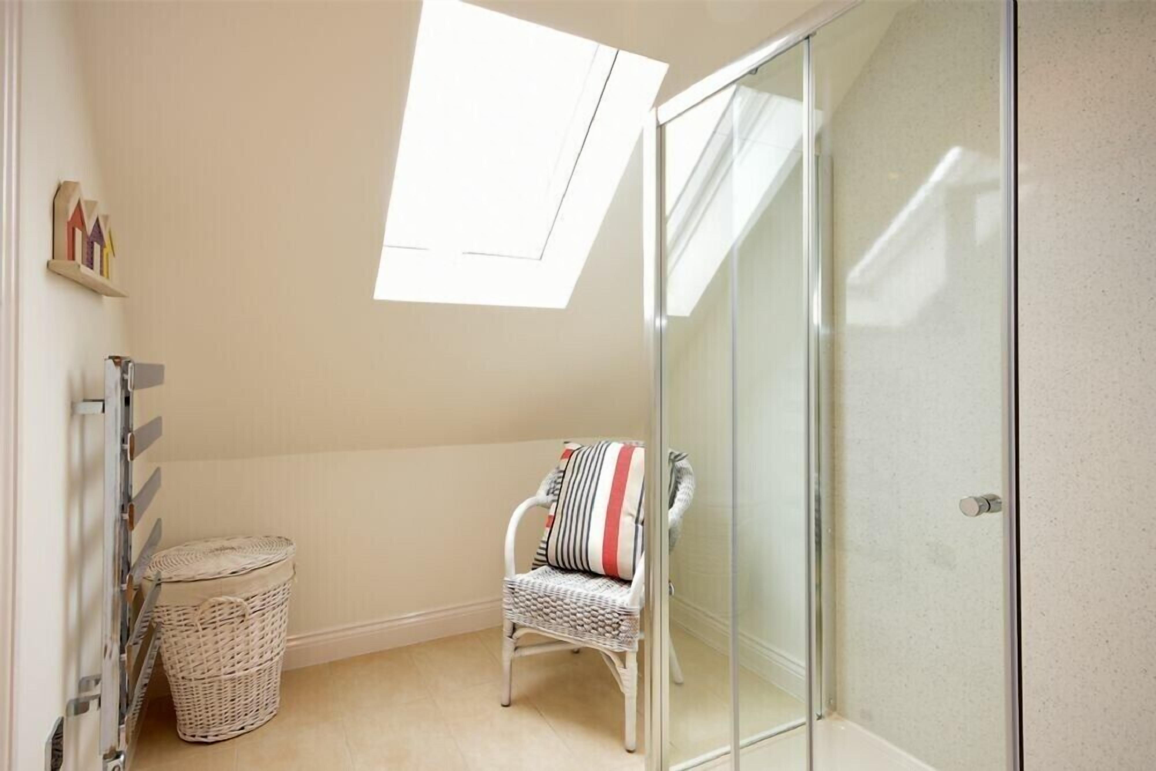 A small, bright room with a skylight, a glass shower enclosure, a wicker chair, and a laundry basket.