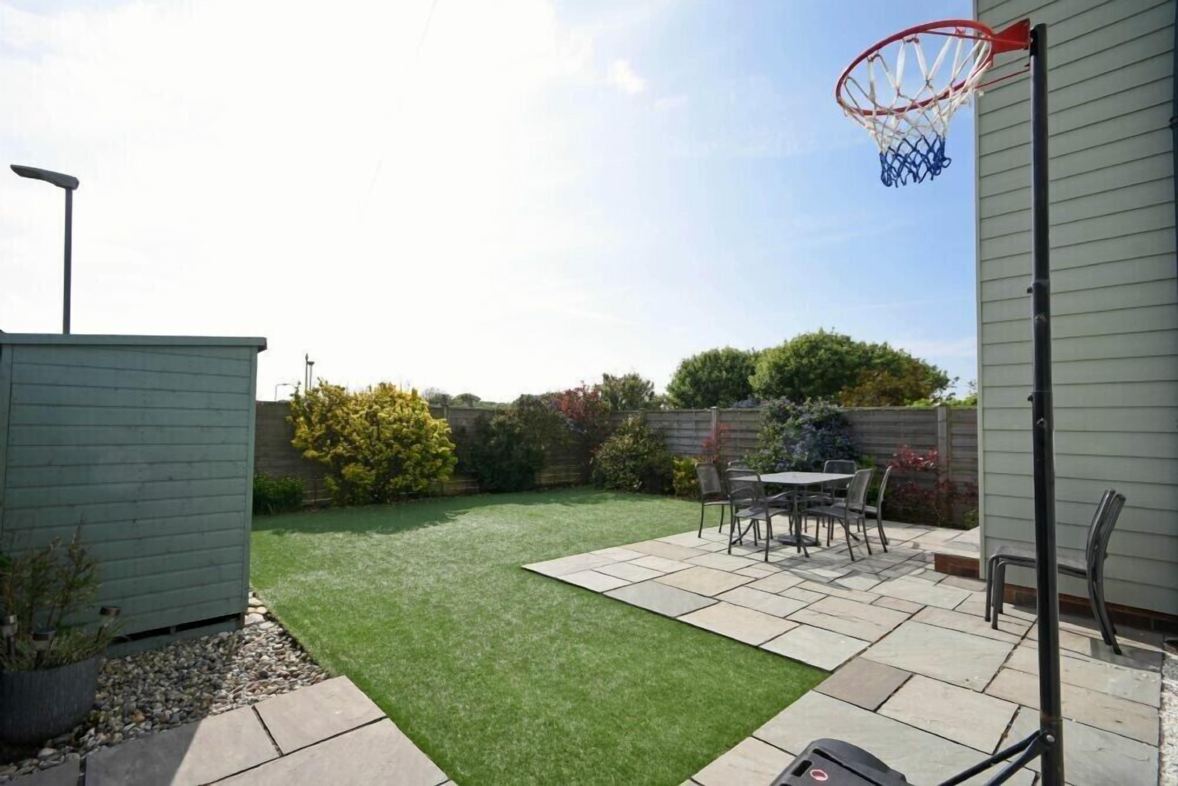 Ein Garten mit Kunstgrasanlage, einem Basketballkorb, einem Patio-Tisch mit Stühlen und einem Geräteschuppen.