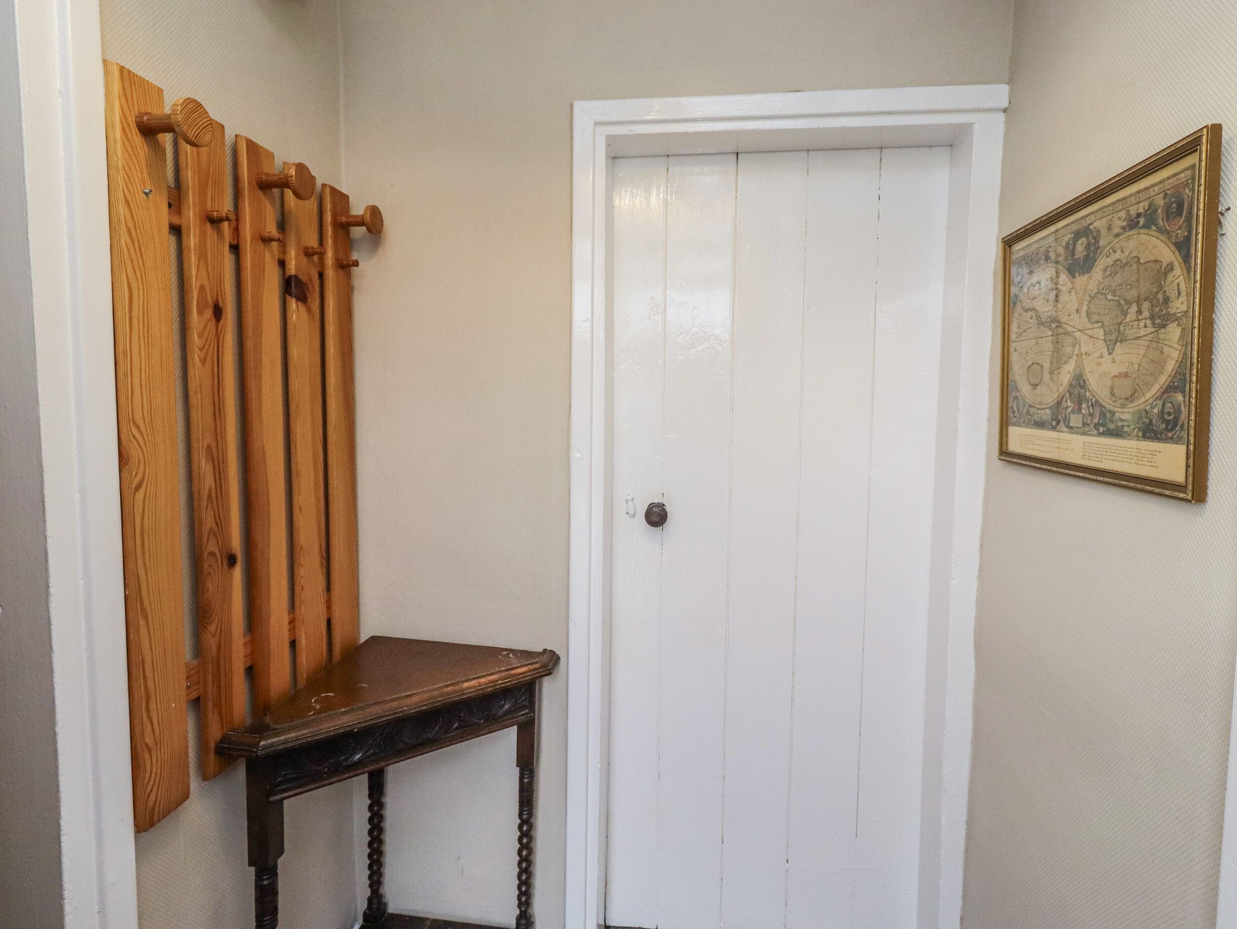 A hallway with a white door, a wooden coat rack, a small table, and a framed map on the wall.