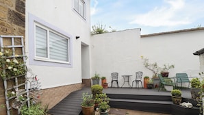 A small patio with a wooden deck, two black metal chairs, a small table, and various potted plants. - Strandline (Alnwick)