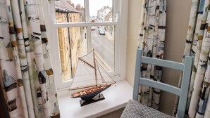 A window with a sailboat model on a windowsill, curtains with a house pattern, and a view of a street with cars and buildings. - Strandline (Alnwick)