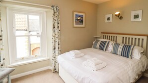 A neatly made bed with white linens and striped pillows, a window with patterned curtains, and a wall with framed pictures. - Strandline (Alnwick)
