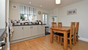A compact kitchen with a dining area, featuring a wooden table and chairs, a window with curtains, and a clock on the wall. - Schooner Court No2 (Alnwick)