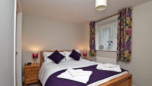 A neatly made bed with a purple bedspread and white linens, a wooden nightstand with a lamp, and a window with floral curtains. - Schooner Court No2 (Alnwick)