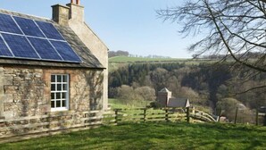 A stone house with a solar panel roof, a wooden fence, and a view of a hilly landscape. - Kirknowe (Hawick)