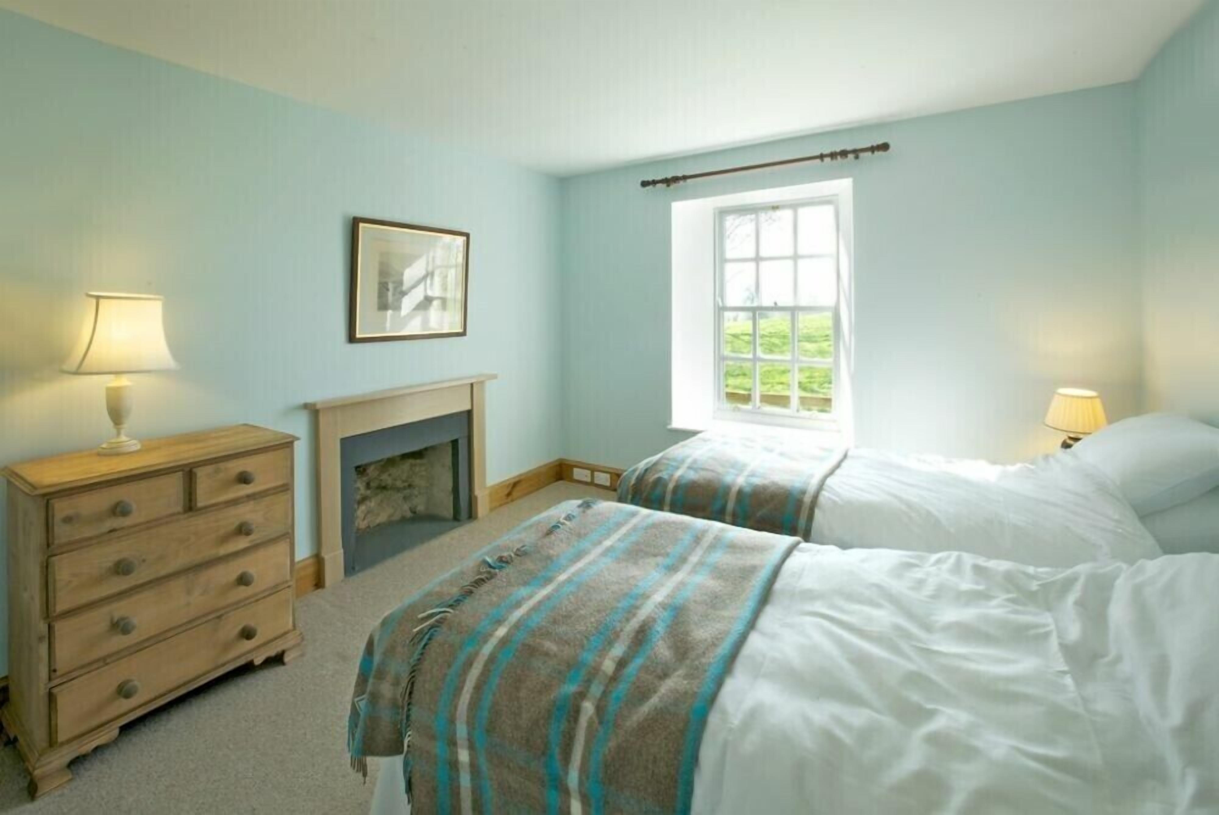 A bedroom with two beds, a dresser, a fireplace, and a window with a view of greenery.