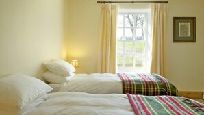 A bedroom with two beds, a window with curtains, a wall-mounted picture, and a bedside lamp. - Kirknowe (Hawick)