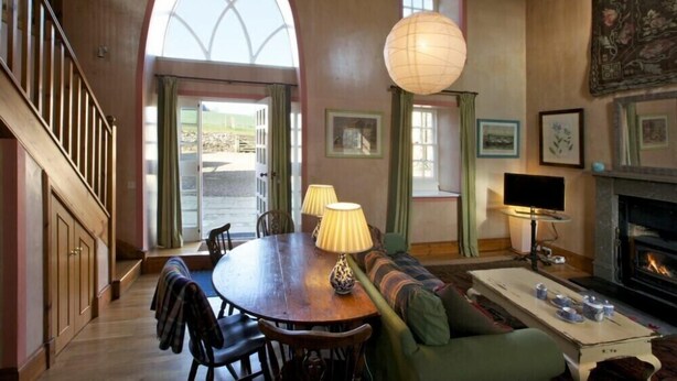 A cozy living room with a wooden staircase, a dining table with chairs, a sofa, a fireplace, and a large arched window. - Kirknowe (Hawick)