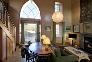 A cozy living room with a wooden staircase, a dining table with chairs, a sofa, a fireplace, and a large arched window. - Kirknowe (Hawick)