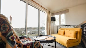 A room with a yellow sofa, a round table with a book, and a view through large windows.