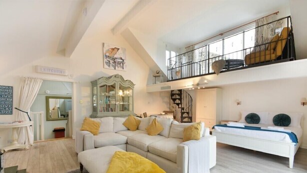 A spacious living room with a white sofa, yellow throw pillows, and a grey rug. A staircase leads to a balcony with a yellow chair.