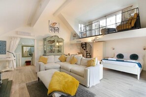 Amplia sala de estar con un sofá blanco, cojines amarillos y una alfombra gris. Una escalera conduce a un balcón con una silla amarilla.