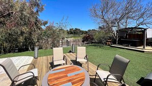 Un patio al aire libre con una mesa de madera redonda y cuatro sillas, rodeado de un césped cuidado y árboles.