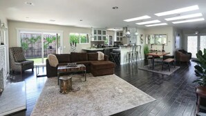 Amplia sala de estar con un sofá seccional grande, una mesa de centro y una alfombra. Hay una zona de cocina con barra y una mesa de comedor. El espacio tiene pisos de madera oscura y ventanas grandes.