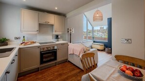 A compact kitchen with modern appliances, a dining area with a table set for two, and a large window offering a view of the city. - Castle View (Bamburgh)
