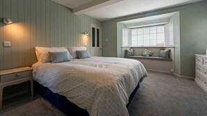 A bedroom with a large bed, a nightstand, a dresser, a window with blinds, and a built-in bench with cushions. - Castle View (Bamburgh)