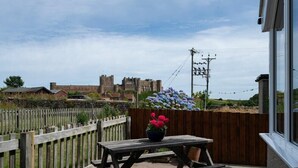 A wooden picnic table with a flower pot, a wooden fence, and a view of a castle-like structure in the background. - Castle View (Bamburgh)