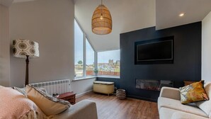 A modern living room with a large window, a fireplace, and a flat-screen TV mounted on the wall. - Castle View (Bamburgh)